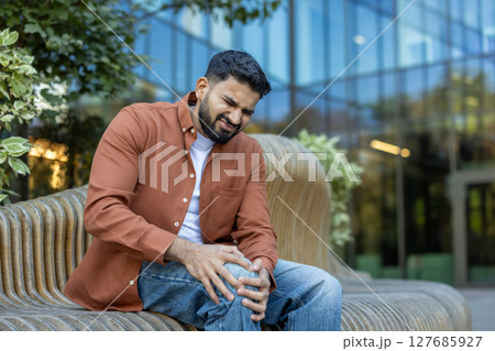 A man is sitting on a bench, visibly experiencing knee pain, his expression showing discomfort and strain. The photo is taken outdoors, during the day. A man is sitting on a bench, visibly experiencing knee pain, his expression showing discomfort and strain. The photo is taken outdoors, during the day. 127685927