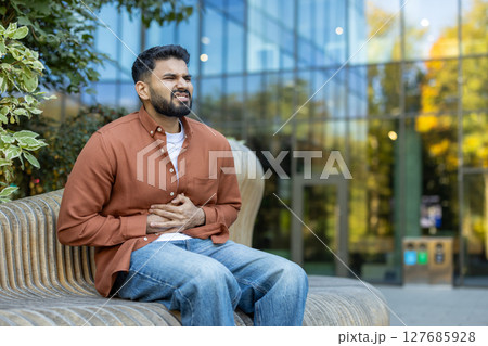 A man, seated outdoors, clutches his stomach, displaying discomfort, suggesting potential illness or pain. The setting includes a bench and a modern building in the background. A man, seated outdoors, clutches his stomach, displaying discomfort, suggesting potential illness or pain. The setting includes a bench and a modern building in the background. 127685928
