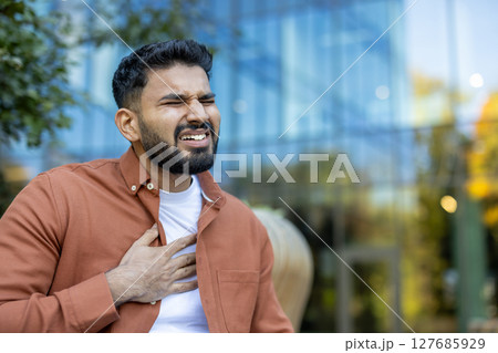 A man clutches his chest in pain, possibly experiencing a heart problem, in an outdoor setting. The background shows a blurred modern building. A man clutches his chest in pain, possibly experiencing a heart problem, in an outdoor setting. The background shows a blurred modern building. 127685929