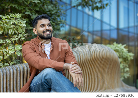A smiling man in a casual outfit sits on a wooden bench, enjoying a sunny day outdoors with a modern building backdrop. A smiling man in a casual outfit sits on a wooden bench, enjoying a sunny day outdoors with a modern building backdrop. 127685936