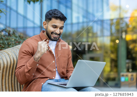 A smiling man sits outdoors with a laptop, clenching his fist in excitement. He appears elated, possibly celebrating a success or good news he received. 127685943