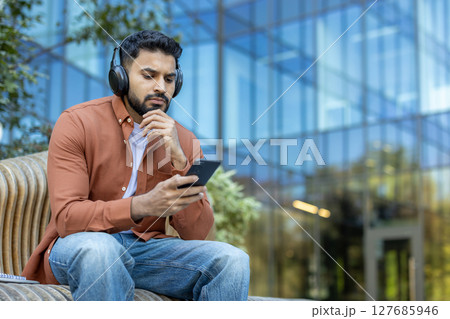 A man wearing headphones sits outside, holding his phone, looking thoughtful with a modern building backdrop. A man wearing headphones sits outside, holding his phone, looking thoughtful with a modern building backdrop. 127685946