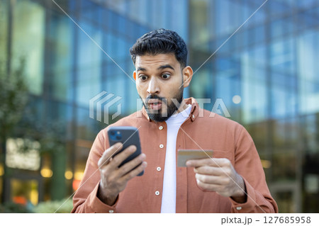 An amazed man looks at his phone while holding a credit card, seemingly surprised by something. An amazed man looks at his phone while holding a credit card, seemingly surprised by something. 127685958