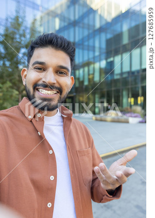 A man smiling and gesturing with his hand during a video call with a modern building in the background. He is friendly 127685959