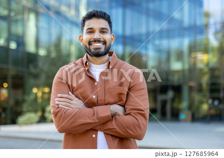 A smiling man stands confidently with arms crossed, posing in front of a modern glass building. The outdoor setting captures a professional and approachable vibe. 127685964