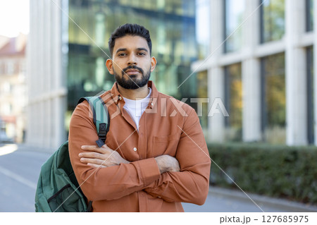 A young, bearded man stands confidently with arms crossed, wearing a button-down shirt, and a backpack. The background features a modern building. 127685975