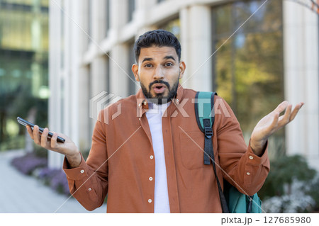 A surprised young man with a backpack and a smartphone looks at the camera with raised hands, outdoors. He seems unsure. 127685980