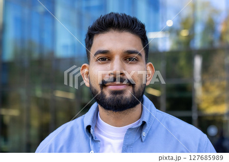 A handsome man with a beard smiles directly at the camera while standing outside. The background shows a modern building with glass windows. A handsome man with a beard smiles directly at the camera while standing outside. The background shows a modern building with glass windows. 127685989