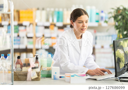 Girl pharmacist waits for visitors near cash register area with computer and greets patients Girl pharmacist waits for visitors near cash register area with computer and greets patients 127686099
