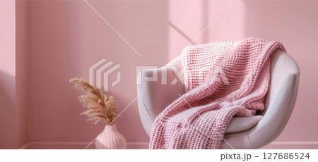 White modern armchair with soft pink knitted blanket and green potted plant on pale pastel background. Minimalist cozy interior design with copy space for text in horizontal layout 127686524