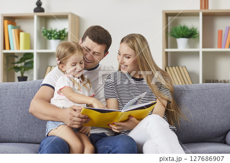 Young happy family on couch reading to their daughter with girl showing book pictures to parents. Young happy family on couch reading to their daughter with girl showing book pictures to parents. 127686907