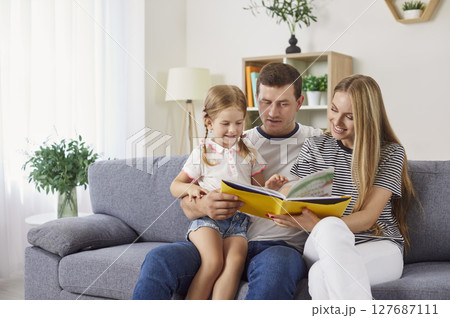 Young family reading book together on couch with father holding daughter and mother showing pictures 127687111