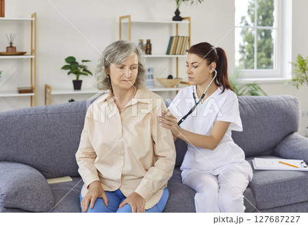Doctor with stethoscope examining woman's breathing and heartbeat visiting senior patient at home 127687227