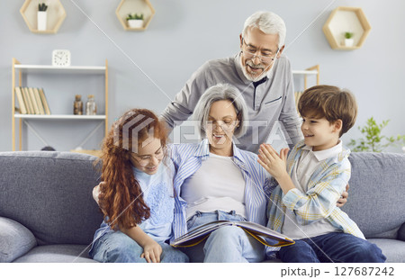 Grandparents reading a book to their grandchildren sitting at sofa at home. 127687242