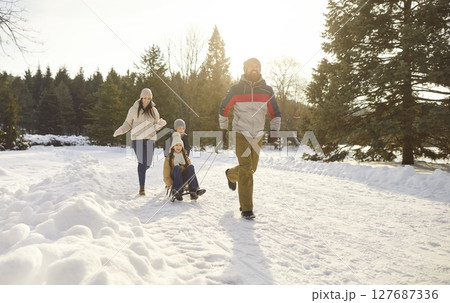 Parents take children on sleds while happily walking along snowy path in winter park. 127687336