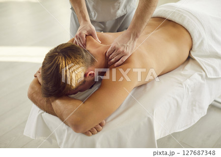 Man client lying on massage table receiving remedial back massage from masseur or physiotherapist 127687348