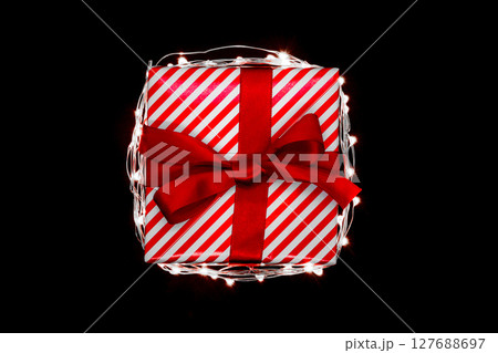 Christmas gift box with red ribbon encircled with illuminated garland and isolated on black background 127688697