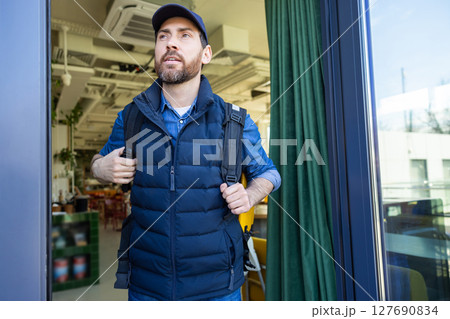Man with delivery bag exiting cafe in blue vest and cap 127690834