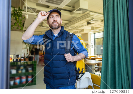 Delivery courier with yellow bag entering modern cafe 127690835
