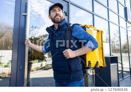 Man with delivery bag exiting cafe in blue vest and cap opening glass door 127690836