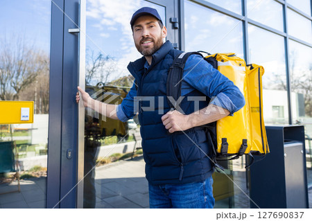 Man with delivery bag exiting cafe in blue vest and cap opening glass door 127690837