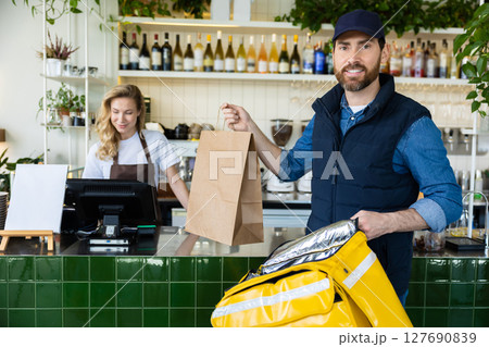 Worker at cafe counter handing order to delivery person in uniform with paper bag 127690839