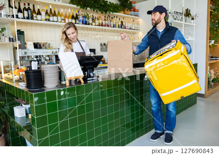 Barista passing delivery order to courier at cafe counter with bag Barista passing delivery order to courier at cafe counter with bag 127690843
