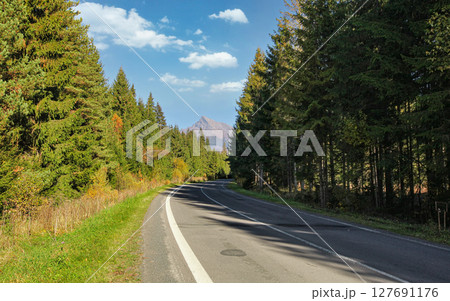 Country asphalt road, coniferous trees on both sides, mount Krivan peak (Slovak symbol)  with blue sky above, in distance 127691176