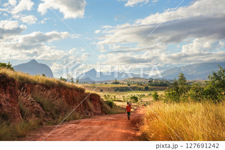 Lone unknown young Malagasy man with basket on his shoulders walking over red dusty road, coming home after working the fields, afternoon sun shines to grass and bushes around, mountains in distance 127691242