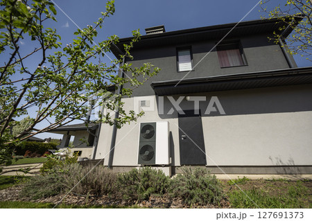 Energy-efficient heat pump unit installed on a modern house exterior surrounded by green yard and landscaping Energy-efficient heat pump unit installed on a modern house exterior surrounded by green yard and landscaping 127691373