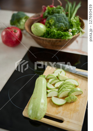 Vertical photo of slices of sliced zucchini are on the cutting board on the kitchen table, a kitchen knife, plate with broccoli, green onion, red radish, lettuce, pomegranate 127693439