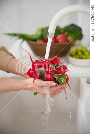 Close up view of a woman washes a bunch of red radishes under the pressure of flowing water in the white kitchen, a basket with tomato, broccoli and onions in the background, grapes 127693449