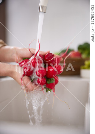 Close up view of a woman washes a bunch of red radishes under the pressure of flowing water in the white kitchen, a basket with tomato, broccoli and onions in the background, grapes Close up view of a woman washes a bunch of red radishes under the pressure of flowing water in the white kitchen, a basket with tomato, broccoli and onions in the background, grapes 127693453