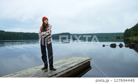 Wide shot, front view of a beautiful young tourist woman or female hiker standing alone on a wooden pier by a forest lake in Finland. She enjoys nature, silence. Concept of mindful travel, eco-tourism Wide shot, front view of a beautiful young tourist woman or female hiker standing alone on a wooden pier by a forest lake in Finland. She enjoys nature, silence. Concept of mindful travel, eco-tourism 127694448