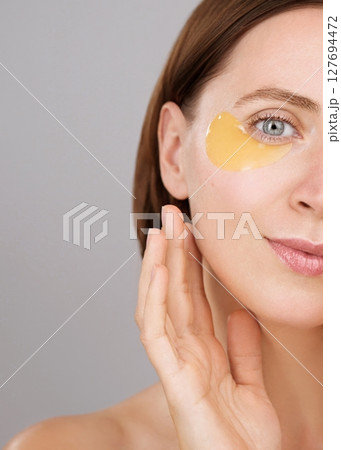 Half face. Eye patch ads concept. Aesthetic woman applying under-eye recovery patches. Skincare. Charming brunette woman in studio on grey background. Cosmetics Beauty routine Dermatology Rejuvenation Half face. Eye patch ads concept. Aesthetic woman applying under-eye recovery patches. Skincare. Charming brunette woman in studio on grey background. Cosmetics Beauty routine Dermatology Rejuvenation 127694472