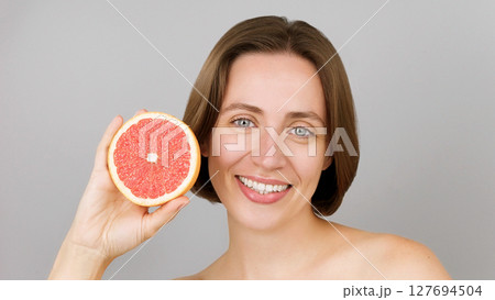 Smiling beautiful Caucasian model playfully holds a grapefruit, showcasing vitamin C benefits. Portrait of a blue-eyed brunette. Commercial eco-friendly beauty skincare, detox nutrition. Grey backdrop 127694504
