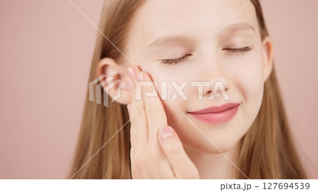 Portrait of smiling cheerful girl doing beauty routine, face massage, applies skin product. Advertisement of self-care products for teenagers. Blonde girl with closed eyes in studio on pink background Portrait of smiling cheerful girl doing beauty routine, face massage, applies skin product. Advertisement of self-care products for teenagers. Blonde girl with closed eyes in studio on pink background 127694539
