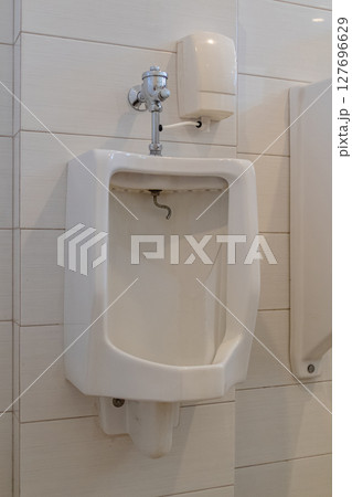 White ceramic urinal with flush valve mounted on tiled wall in public restroom White ceramic urinal with flush valve mounted on tiled wall in public restroom 127696629