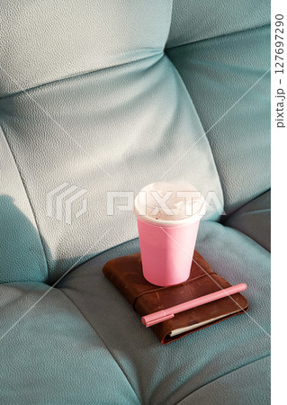 Pink Coffee Cup and Brown Notebook with Pink Pen on a Blue Couch in Bright Sunlight. Pink Coffee Cup and Brown Notebook with Pink Pen on a Blue Couch in Bright Sunlight. 127697290