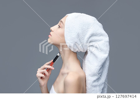 Young caucasian female applying makeup with brush on neck wrapped in towel Young caucasian female applying makeup with brush on neck wrapped in towel 127697414