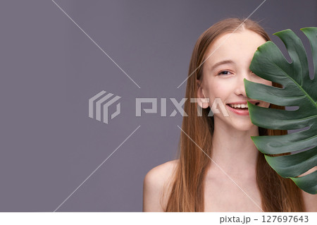 Young caucasian female smiling with monstera leaf on gray background 127697643