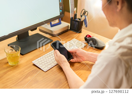 パソコン作業の合間に携帯電話をチェックする50代男性 パソコン作業の合間に携帯電話をチェックする50代男性 127698112