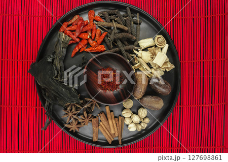 Mix composition of various exotic dried spices in black round bowl on red bamboo mat 127698861