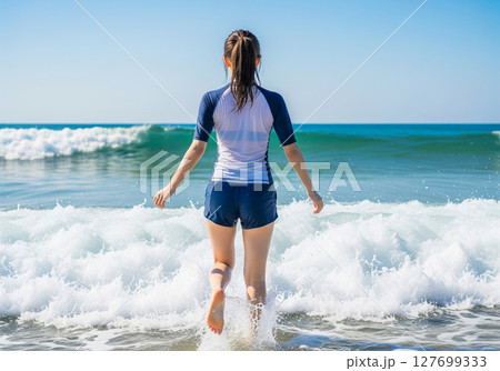波がある海に足を踏み入れる、ラッシュガードを着たポニーテールの女性の後ろ姿 127699333
