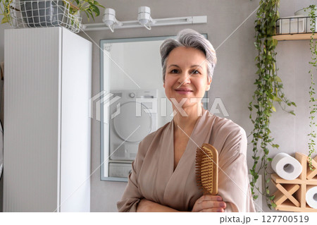 Smiling senior woman grooming her hair in natural light bathroom with wooden brush and plants 127700519