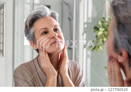 Mature woman examining her face in mirror during morning skincare routine at home Mature woman examining her face in mirror during morning skincare routine at home 127700520