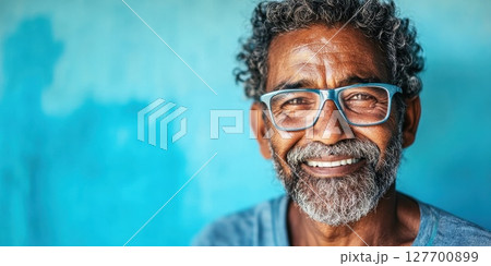 Elderly man with glasses smiles warmly against a textured blue wall in a lively urban setting during the daytime, showcasing joy and wisdom Elderly man with glasses smiles warmly against a textured blue wall in a lively urban setting during the daytime, showcasing joy and wisdom 127700899