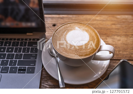 Close-up of Hot coffee latte with latte art milk foam in cup mug and laptop computer and smartphone on wood desk office desk in coffee shop at the cafe,during business work concept Close-up of Hot coffee latte with latte art milk foam in cup mug and laptop computer and smartphone on wood desk office desk in coffee shop at the cafe,during business work concept 127701572