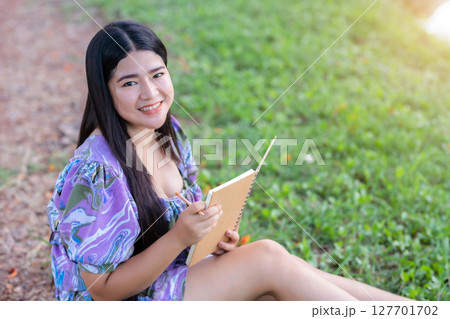 Happy Relaxing Portrait of freelancer asian woman Wear purple dress while working holding diary book writing note while sitting on green grass lawn beside a reservoir at the city park outdoors. 127701702