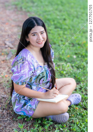 Happy Relaxing Portrait of freelancer asian woman Wear purple dress while working holding diary book writing note while sitting on green grass lawn beside a reservoir at the city park outdoors. 127701705